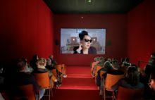 Red auditorium of people watching sunglass wearing woman on cinema screen
