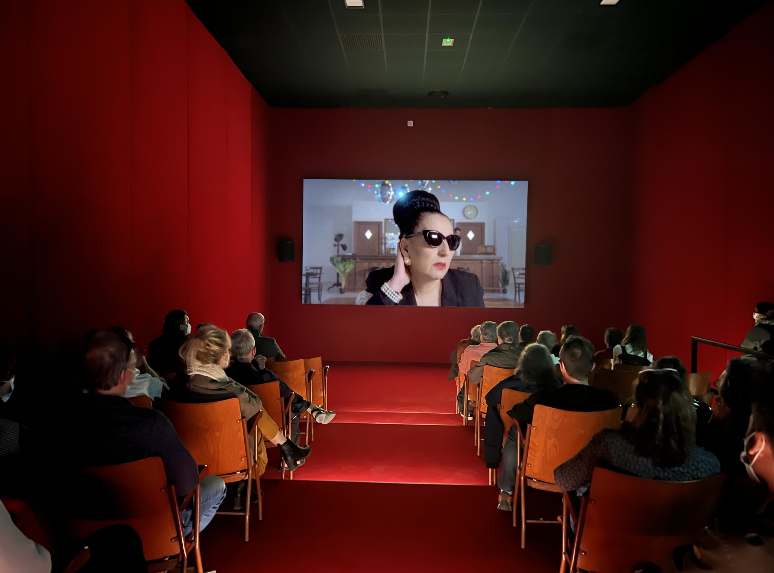 Red auditorium of people watching sunglass wearing woman on cinema screen