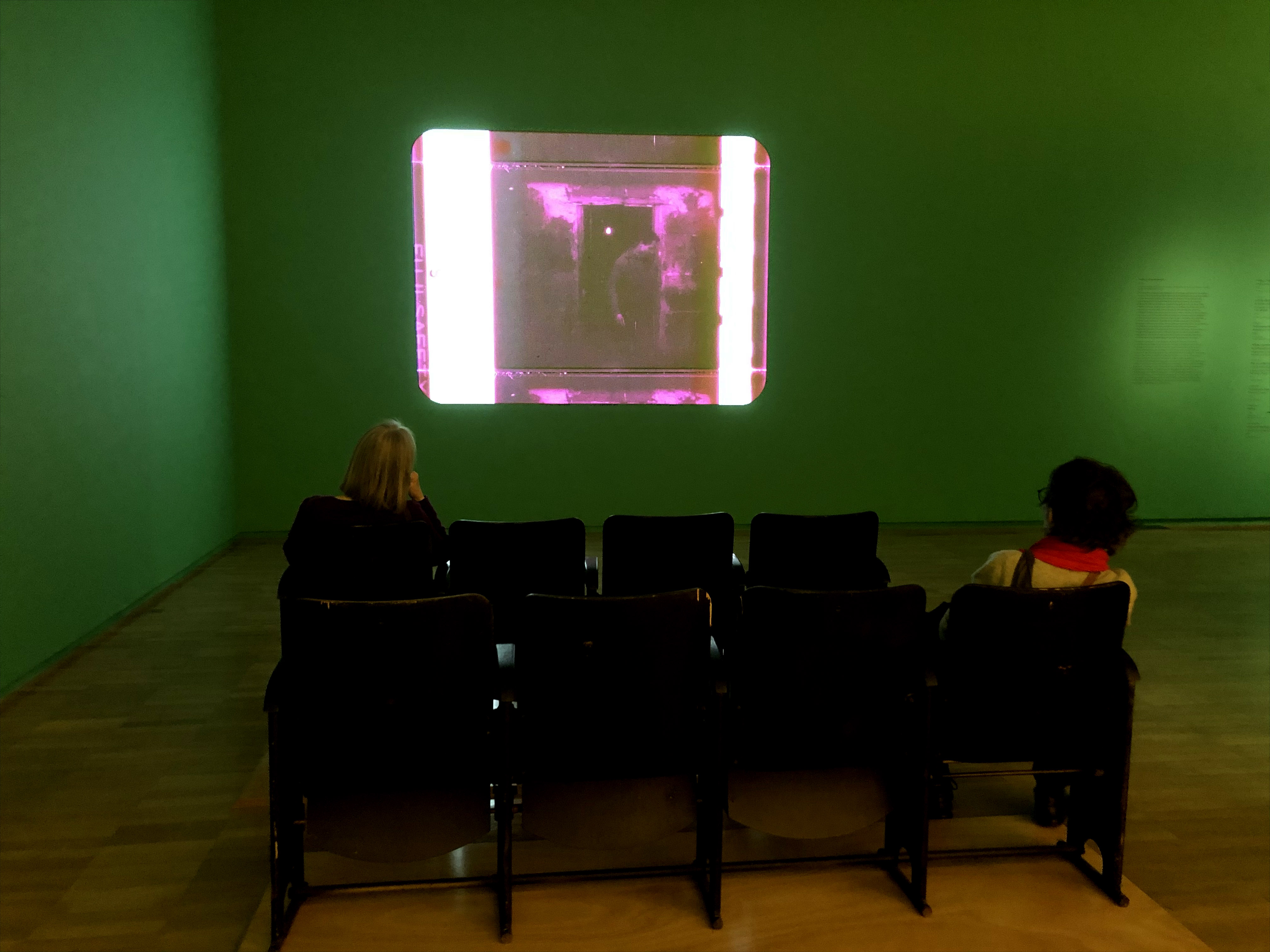 two people sit apart on banks of seats viewing purple tinged projected image on wall