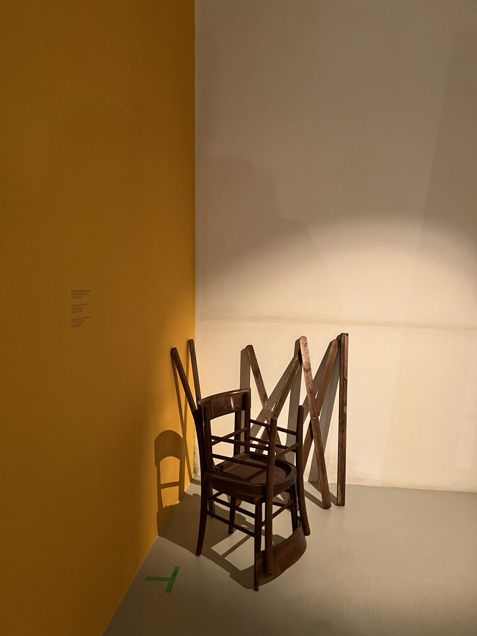 Spot lit in a corner: 2 dark wooden chairs, one stored upside down on other, and dark wood collapsible frame