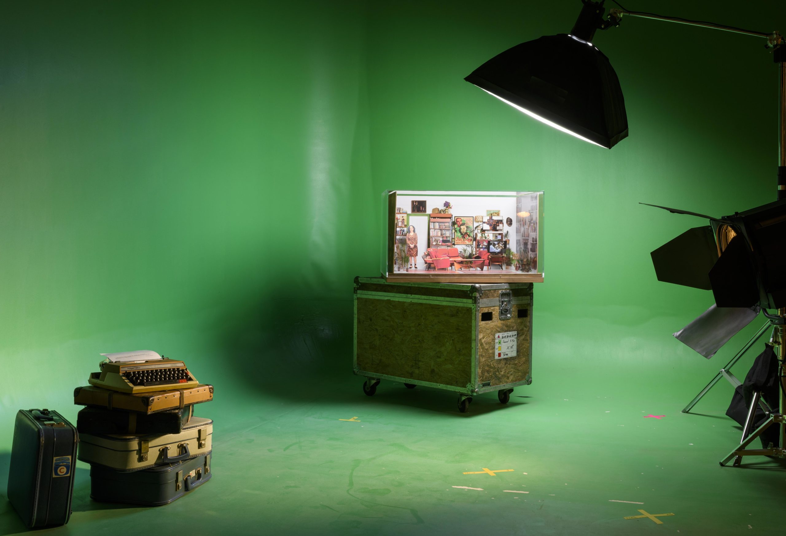 foreground has stack of old suitcases and typewriters while in background apicture stands on wheeled flight case against a greenscreen background - the scene lit by large film set lights