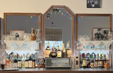 Bar counter - 3 mirrors behind shelves of glasses and many wine & spirits bottles on counter. Centrally on counter is vintage radio